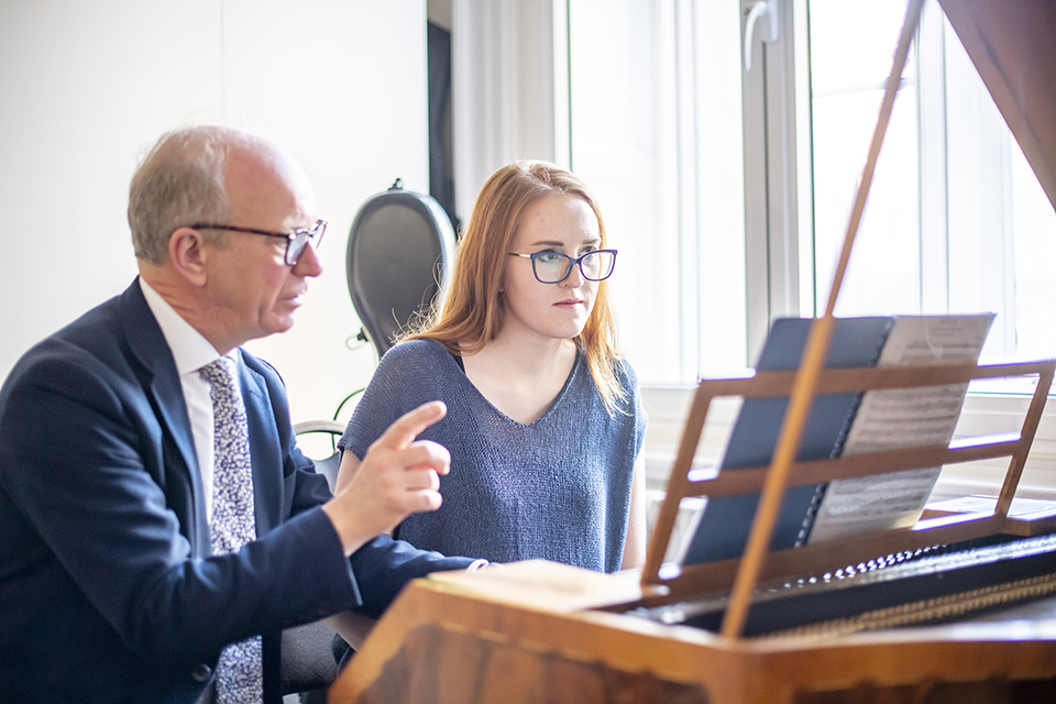 A male professor, with fair hair and glasses, helping educate a female student, playing the harpsichord, in a white, naturally lit room.